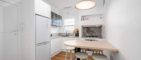 Une cuisine compacte avec des meubles blancs, un four encastré, un évier et une table à manger en bois avec des tabourets.