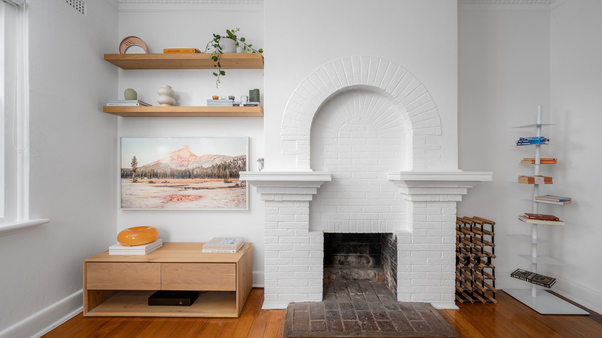 A modern living room with a white brick fireplace, wooden shelves, a painting, and a bookcase.