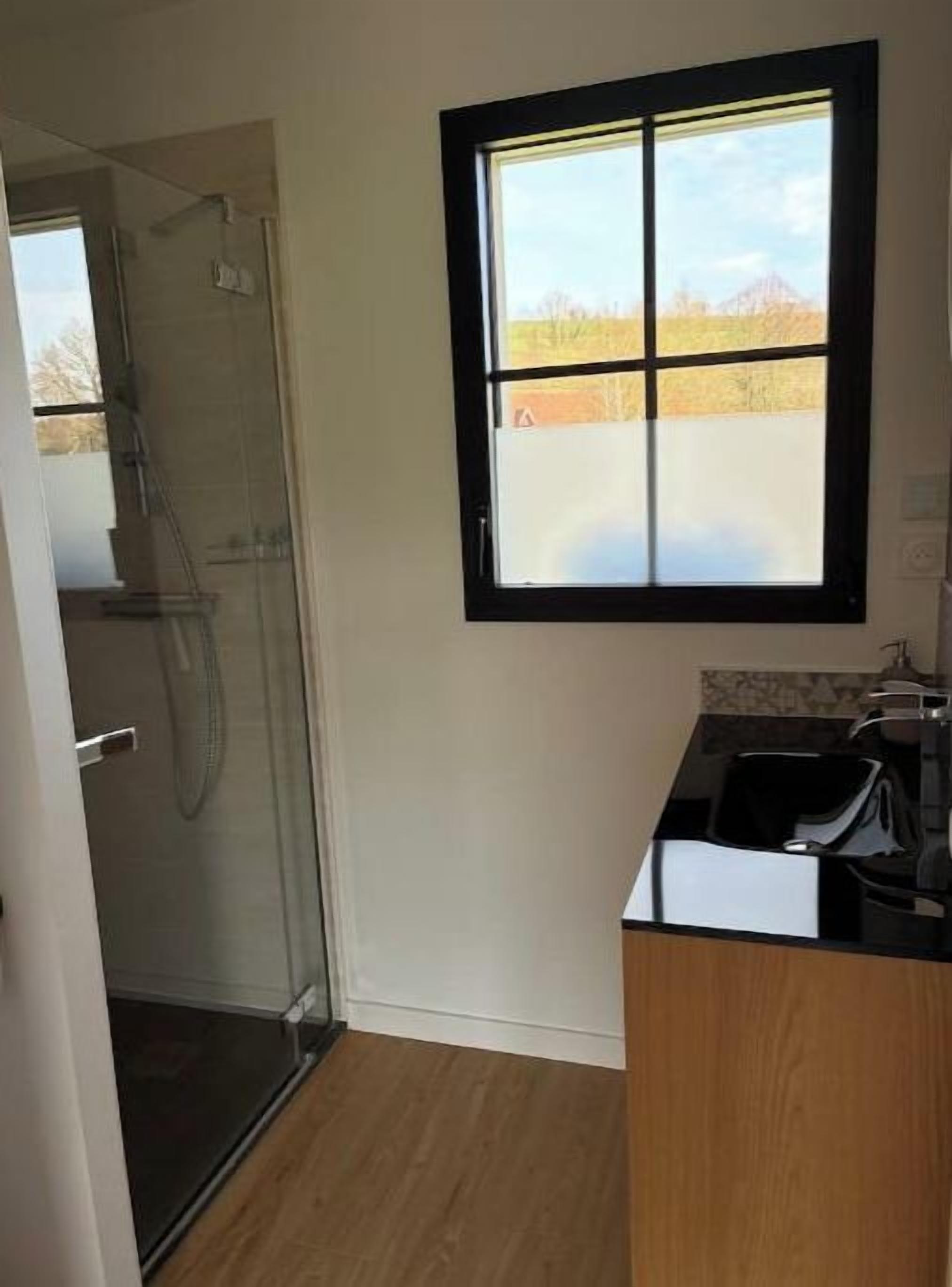 A bathroom with a glass shower enclosure, a window with a black frame, and a wooden floor.