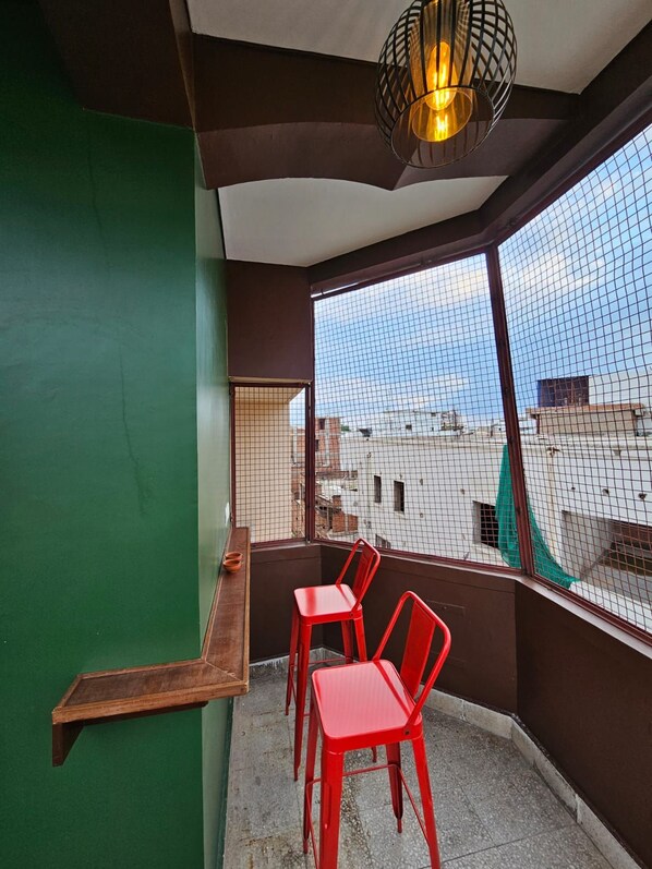 Ein Balkon mit Blick auf die Stadtlandschaft, ein roter Stuhl, ein Holzregal und eine hängende Leuchte.