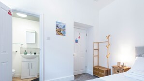 Quarto com cama, cabideiro de madeira, banheiro com pia e armários, e porta com placa de 'Ação contra incêndio'.