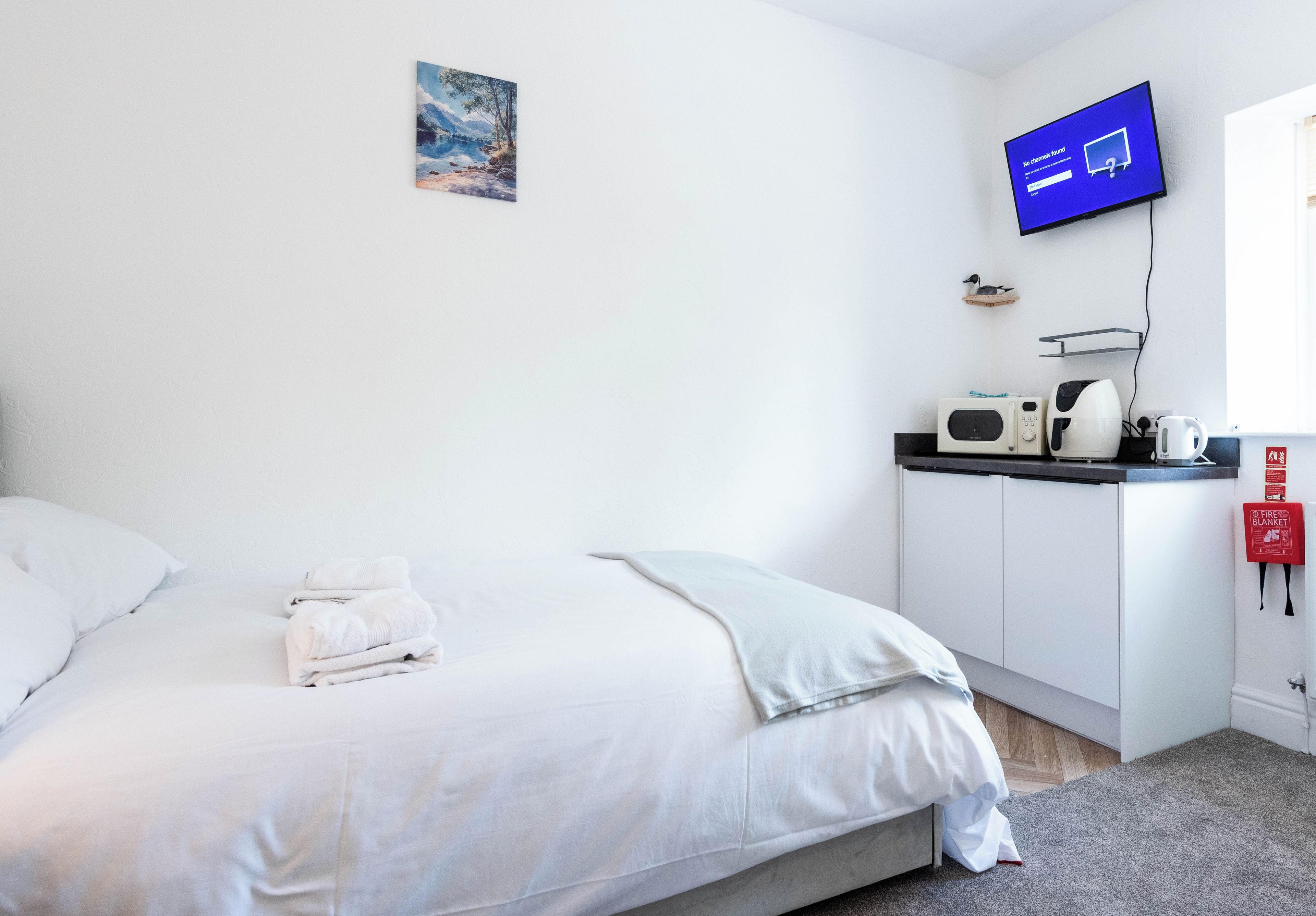 A hotel room with a bed, a flat-screen TV, a microwave, a kettle, and a fire blanket.