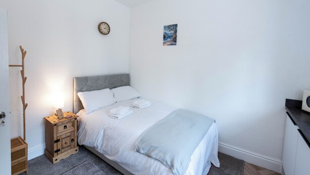 Quarto com cama, mesa de cabeceira com abajur, cabideiro de madeira, relógio e um pequeno quadro na parede.