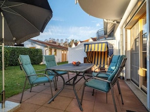 Een terras buiten met een tafel en vier stoelen, een gestreept parasol, en een overdekte zithoek.