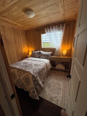 Une pièce en bois avec un lit, deux lampes de chevet, une fenêtre avec des rideaux légers et une porte.