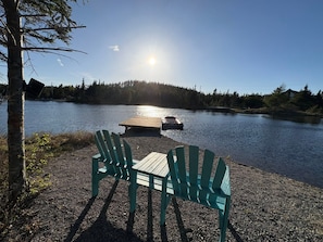 Un cadre idyllique au bord du lac, avec une jetée en bois, deux fauteuils Adirondack verts et une vue dégagée sur le lac et les collines boisées.