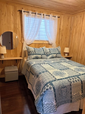 A bedroom with a wooden paneled wall, a bed with a patterned quilt, two bedside lamps, a round mirror, and a window with sheer curtains.