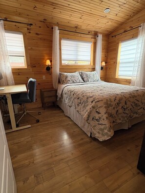 Un dormitori d'una cabana de fusta, amb un llit, escriptori, cadira i dues finestres amb cortines.