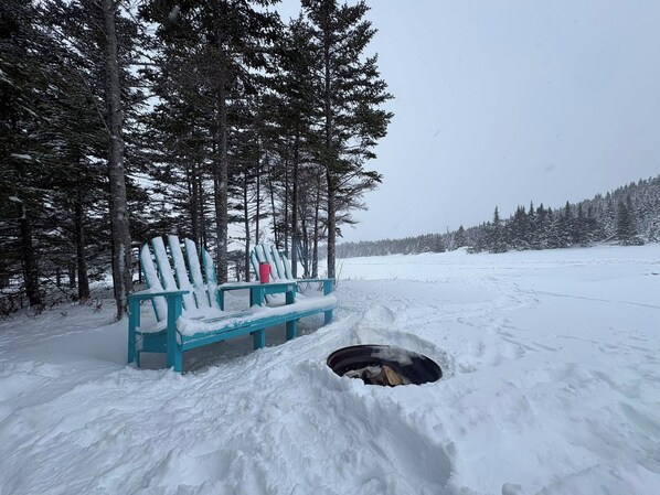 A bench in a snowy forest with a fire pit in the foreground.