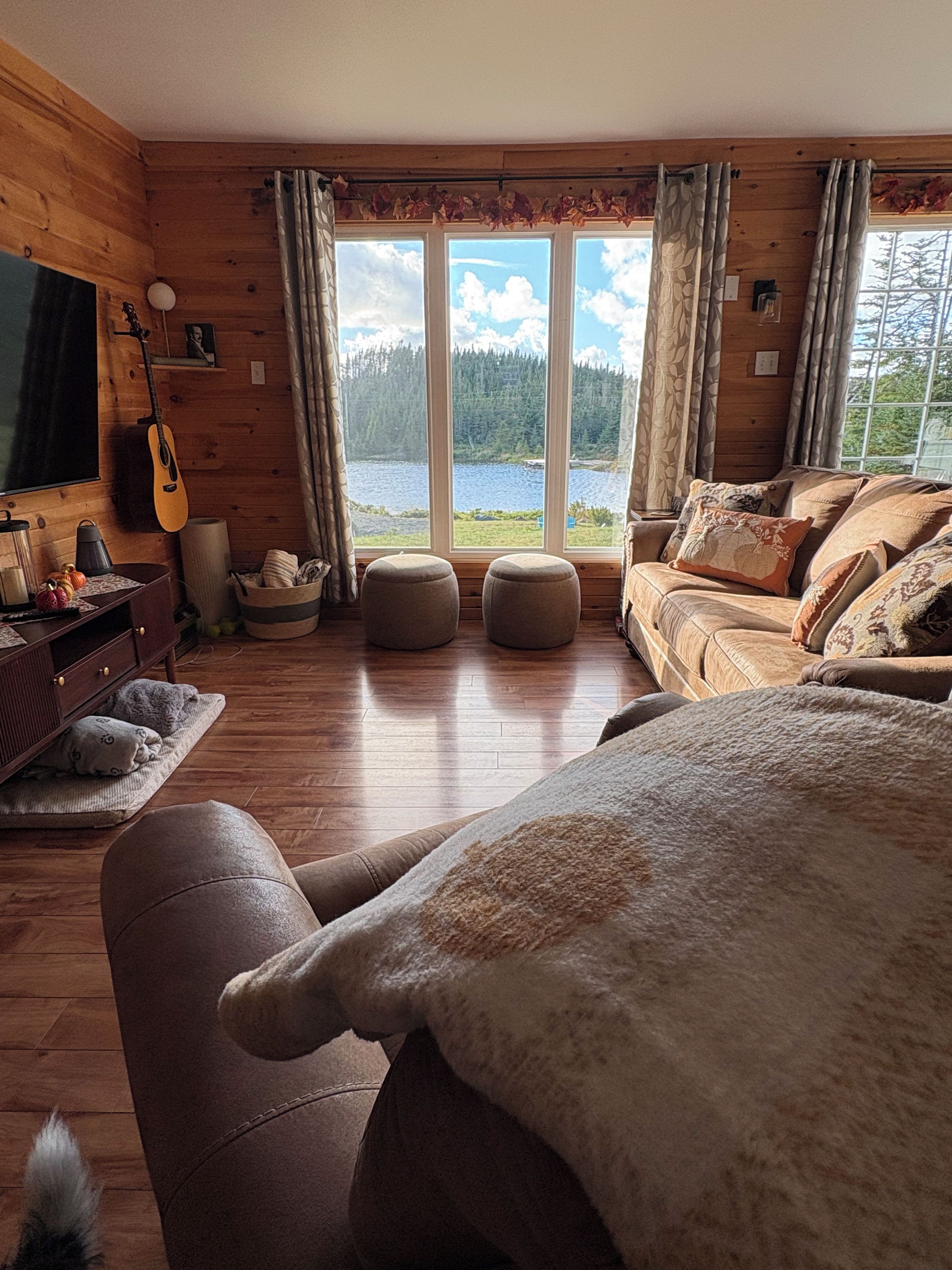 A cozy room with a wooden interior, a large window with a view of a lake, a sofa, ottomans, and a guitar.