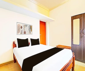 A hotel room with a bed, a wooden nightstand, a door, and a wall-mounted artwork. - Deluxe KS Comfort (Chidambaram)