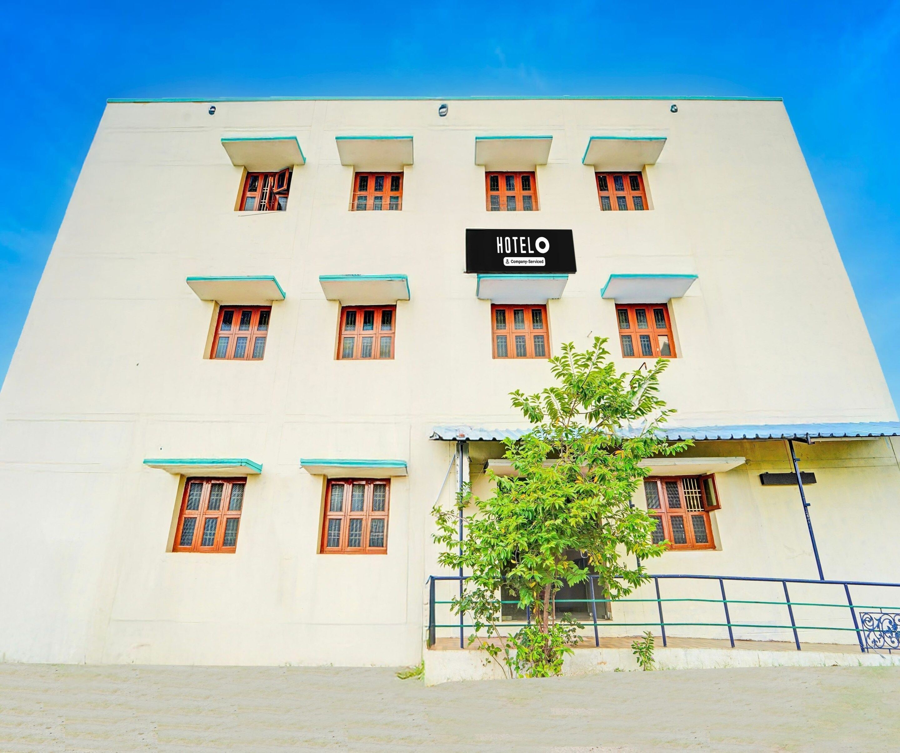A hotel building with a sign that reads 'HOTEL' and a company-serviced logo. There are multiple windows with wooden frames and a tree in front.