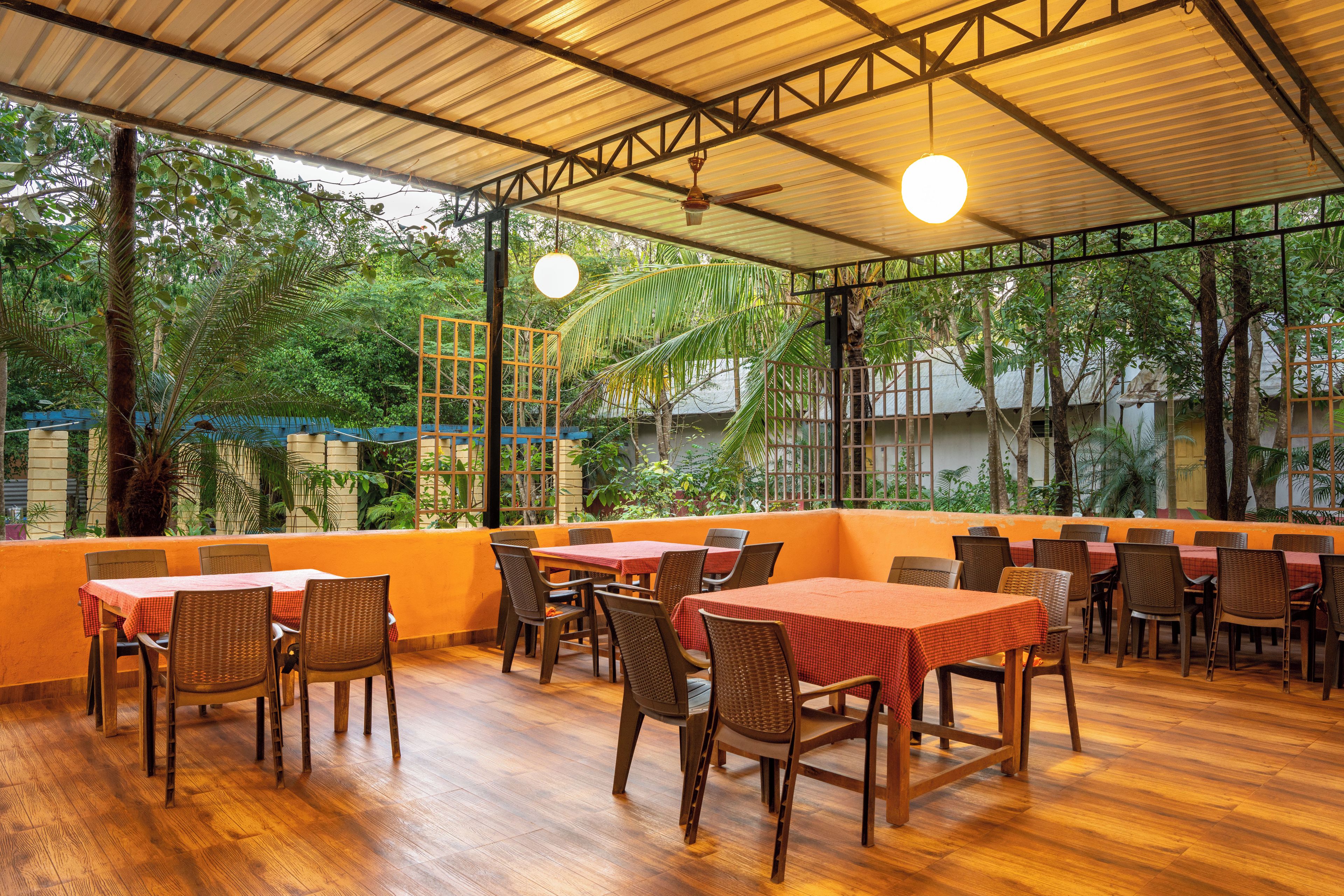 An outdoor restaurant with wooden tables and chairs, surrounded by greenery and a metal roof structure.