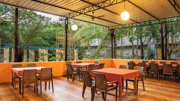 An outdoor restaurant with wooden tables and chairs, surrounded by greenery and a metal roof structure.