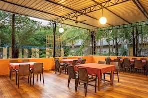 Un restaurante al aire libre con mesas y sillas de madera, rodeado de follaje y una estructura de techo metálico.
