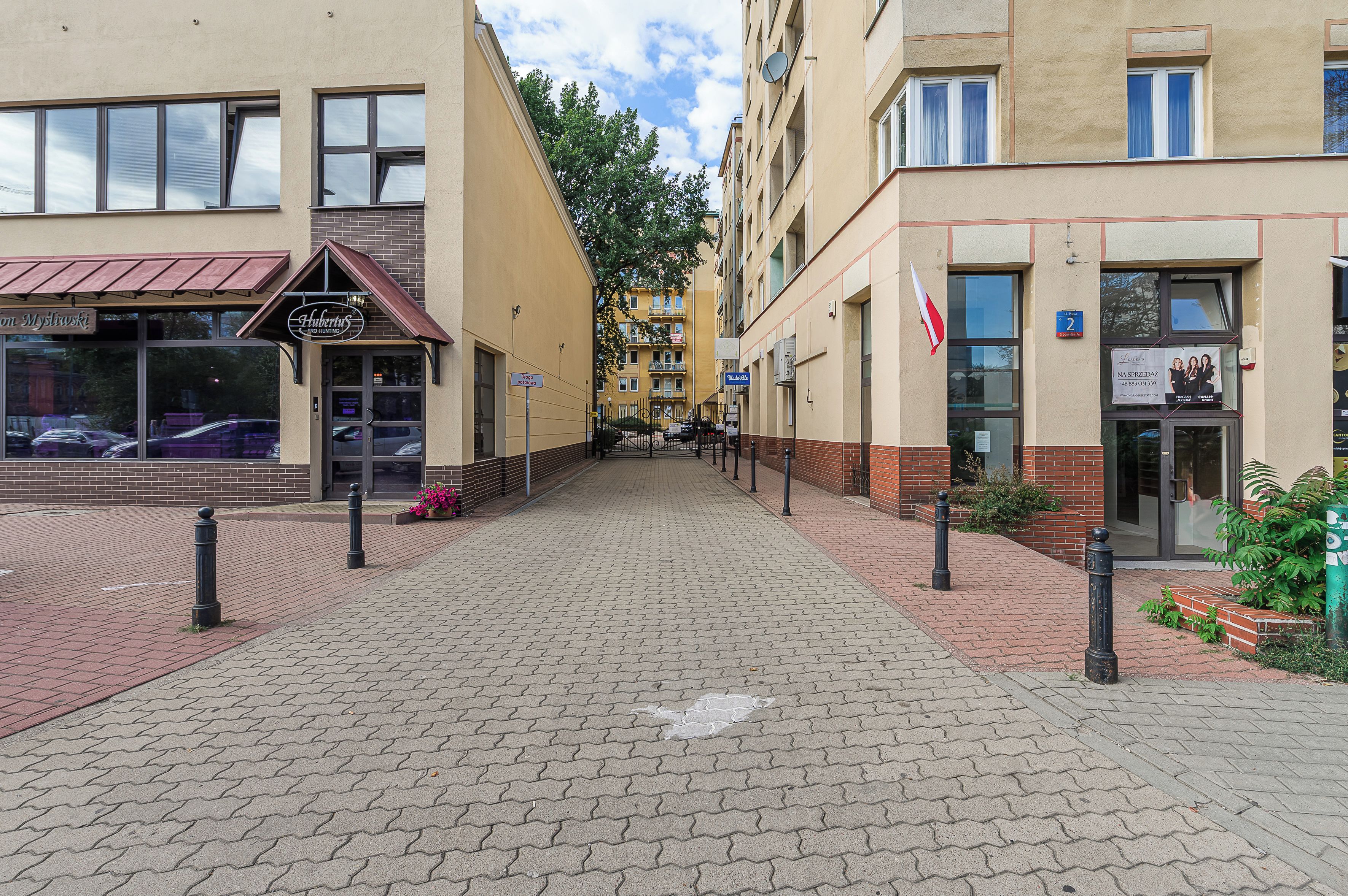 Un chemin pavé mène à un bâtiment dont la façade donne sur la rue, avec une vitrine sur la gauche et un drapeau sur la droite.
