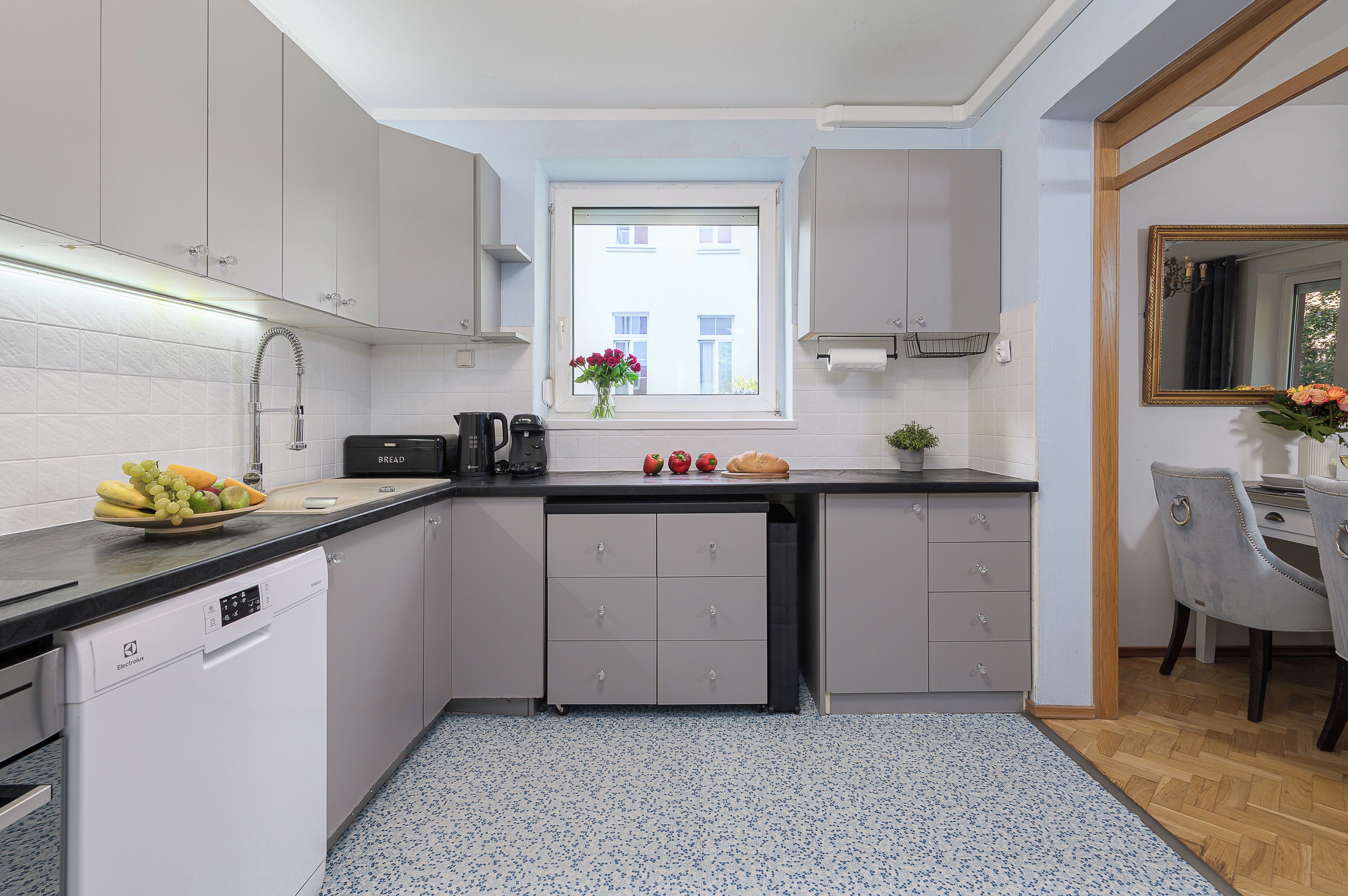 A modern kitchen with grey cabinets, a black countertop, a built-in dishwasher, and a window with a view of buildings.