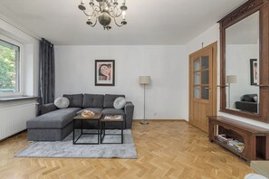 A modern living room with a sectional sofa, a small coffee table, a floor lamp, a large mirror, and a wooden cabinet with books. - Lumina 3-room Apt w/ Balcony I Center (Warszawa)