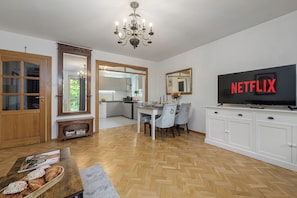 A modern living room with a television displaying 'NETFLIX,' a dining area with a table set for two, and a kitchen in the background. - Lumina 3-room Apt w/ Balcony I Center (Warszawa)