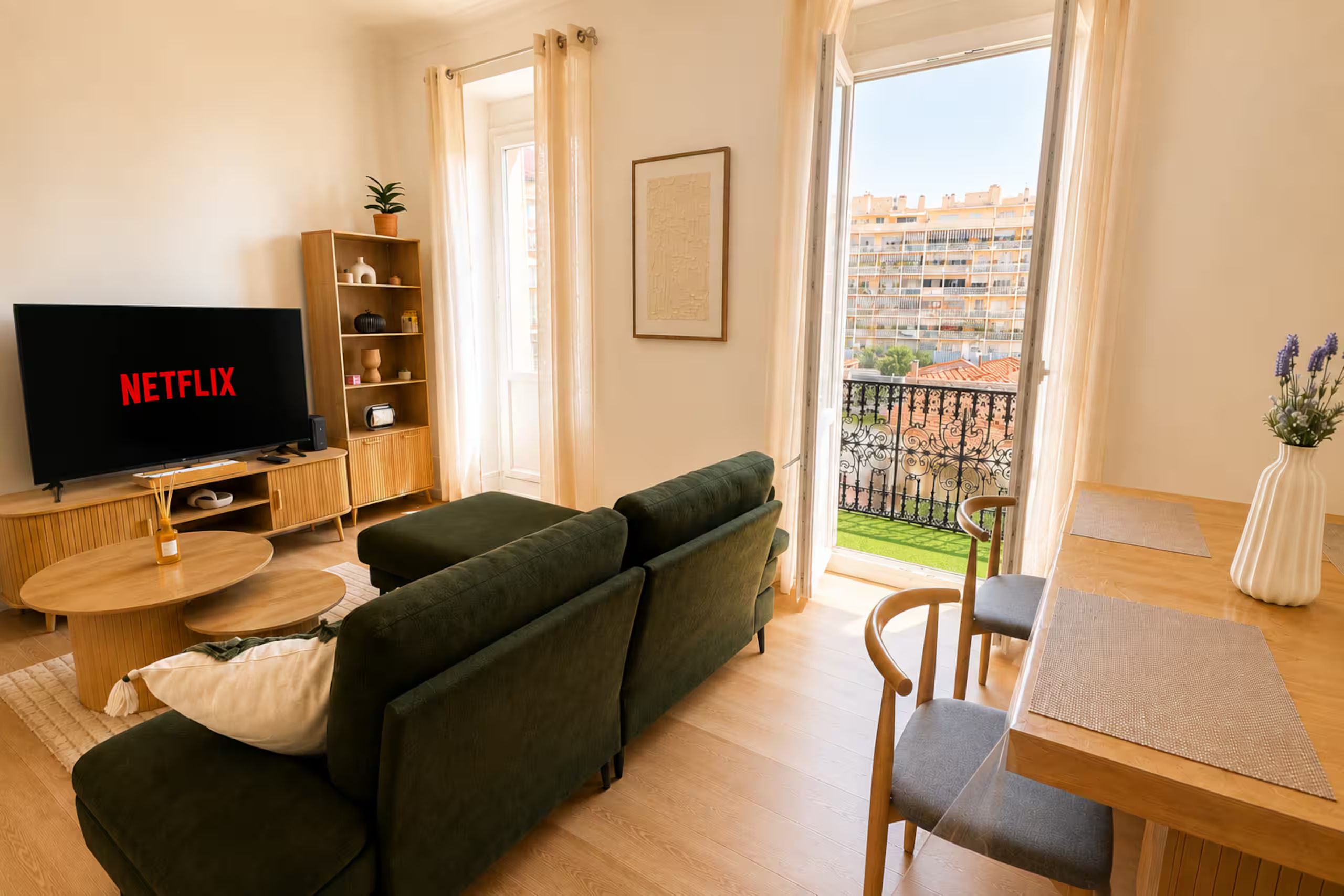 Un salon moderne avec un canapé vert, une table basse en bois, un téléviseur dont l’écran affiche le logo « NETFLIX », une bibliothèque et un balcon offrant une vue sur les immeubles.