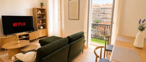 Una sala moderna con un sofá verde, una mesa de centro de madera, un televisor mostrando 'NETFLIX', una estantería y un balcón con vista a edificios.