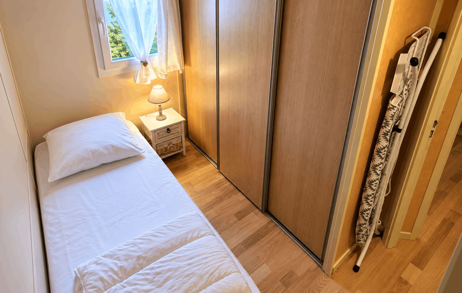 A bedroom with a bed, a nightstand with a lamp, a wardrobe, and a window with curtains.
