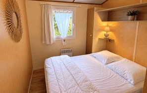 A single bed with a white duvet and pillows, a bedside lamp with a heart-shaped lampshade, a window with sheer curtains, and a wall-mounted mirror.