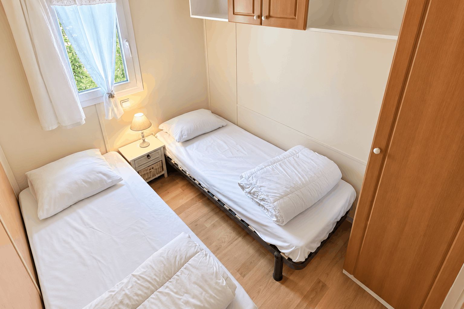 A compact bedroom with two single beds, a bedside table with a lamp, and a window with curtains.