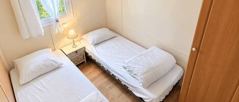 A compact bedroom with two single beds, a bedside table with a lamp, and a window with curtains.