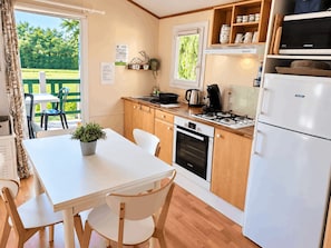 A compact kitchen with a stove, oven, and refrigerator, a dining table with chairs, and a window overlooking greenery.
