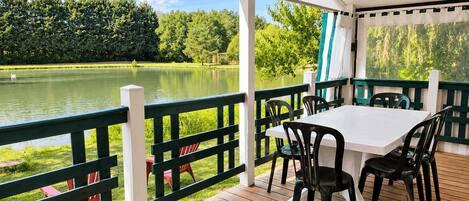 Un espacio exterior techado con una mesa y sillas, con vista a un lago y rodeado de vegetación.