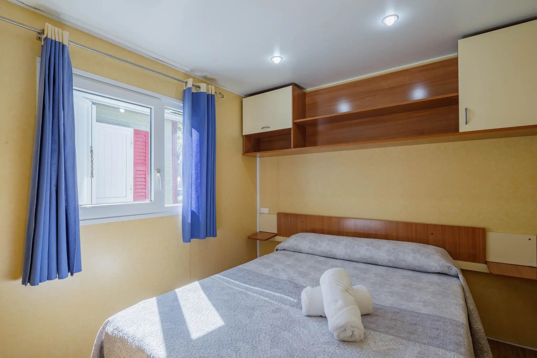Un dormitorio con cama, ventana con cortinas, mesita de noche y espacio de almacenamiento integrado.
