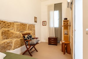Una habitació amb una paret de pedra, una cadira de fusta amb coixí, una taula de fusta petita i una prestatgeria amb penjadors.