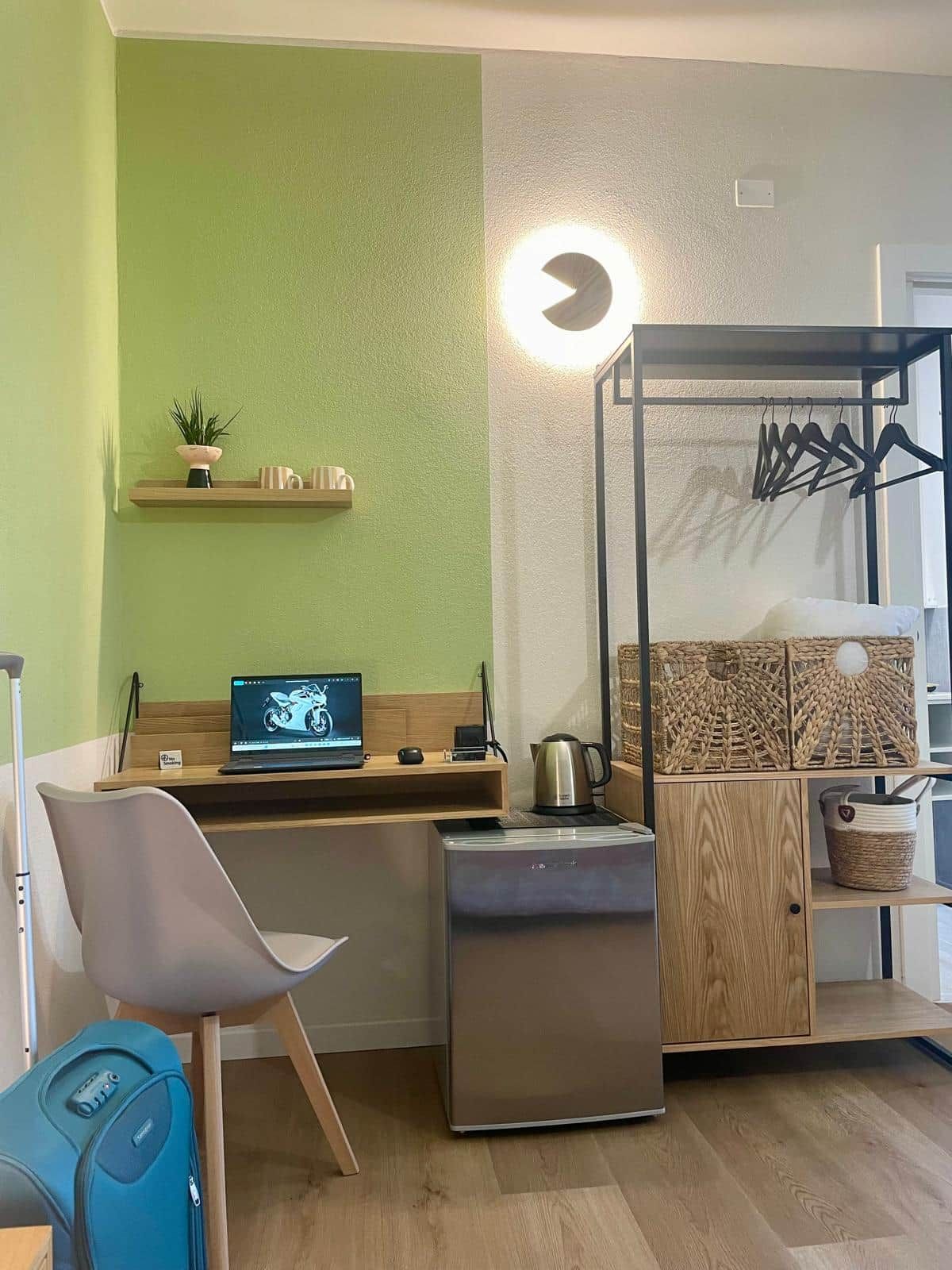 Ein kleines Zimmer mit einem Laptop auf einem Schreibtisch, einem kleinen Kühlschrank, einem Stuhl, einem Koffer und einem Wandregal mit Pflanze und Lampe.