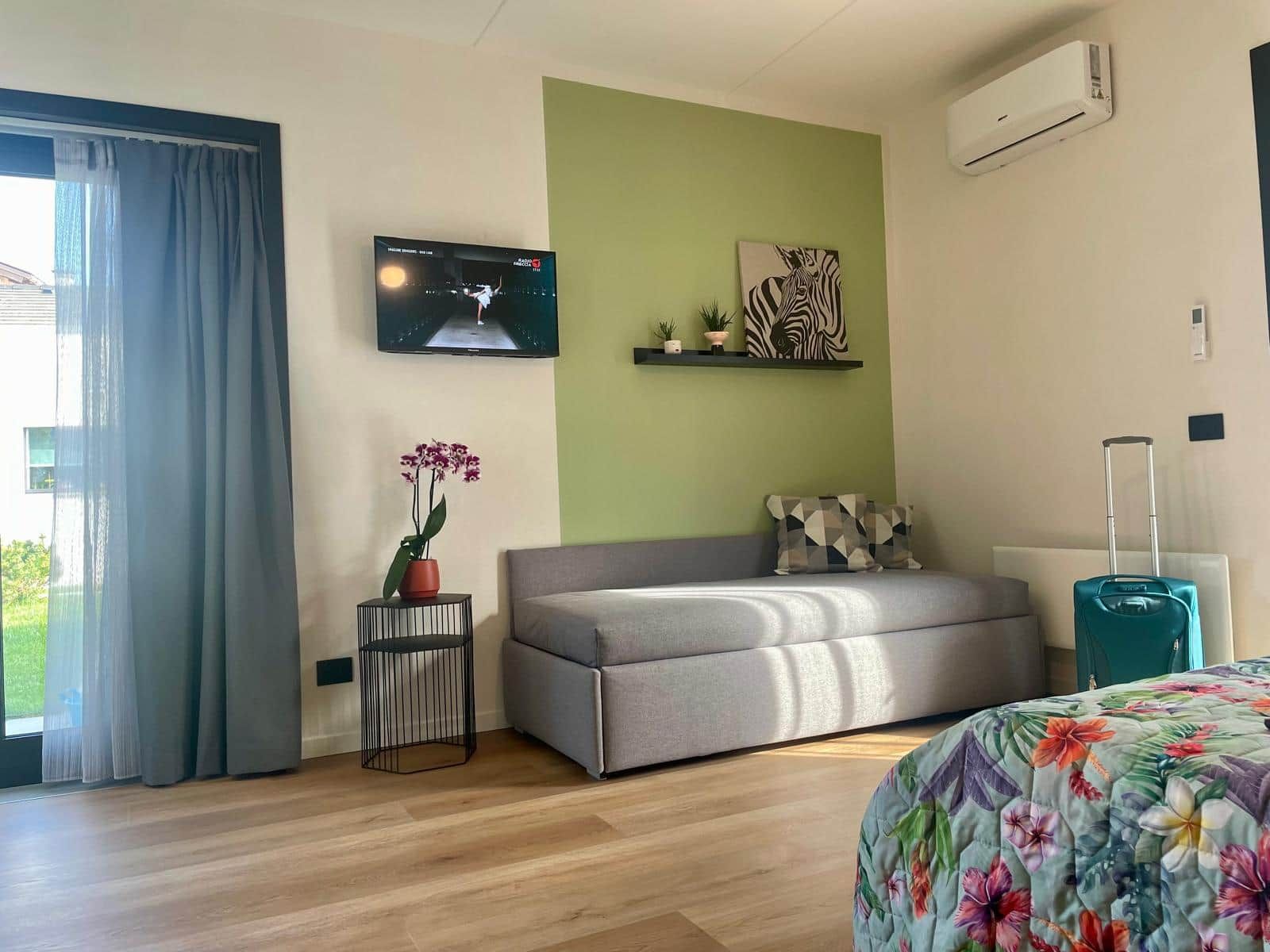 Une chambre d’hôtel moderne avec un canapé gris, un téléviseur à écran plat fixé au mur, une œuvre d’art à motifs zébrés, une petite plante en pot et une valise de couleur turquoise.