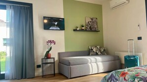 Een hotelkamer met een bank, een bed met bloemenbedekking, een televisie aan de muur, een klein tafeltje met een bloempot en een koffer.
