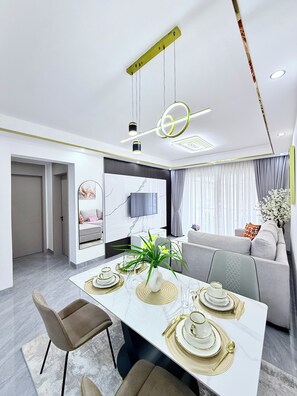 Un comedor moderno con una mesa blanca preparada para cuatro personas, un sofá y un televisor al fondo.