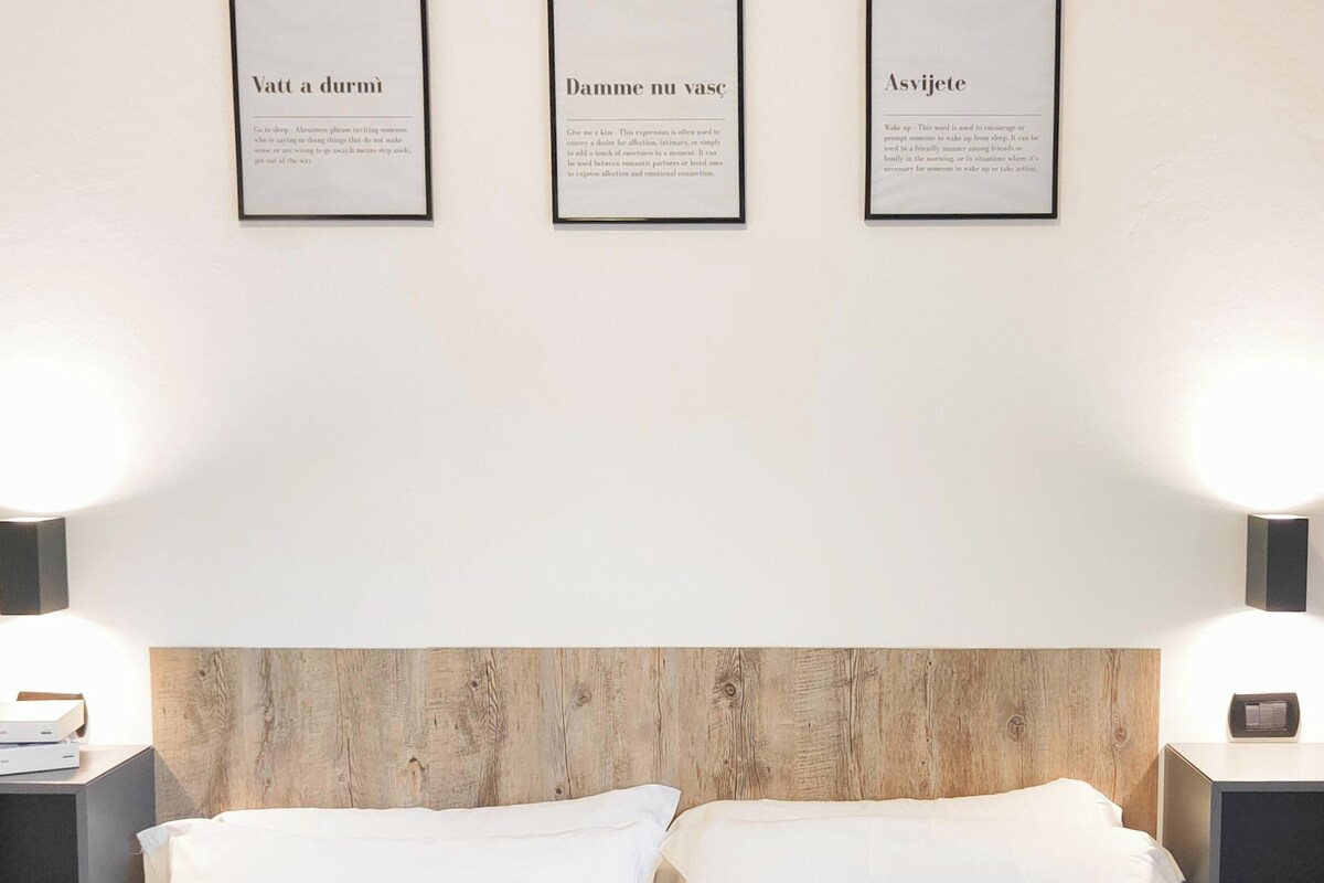A hotel room with a wooden headboard, two bedside lamps, a nightstand with books, and three framed signs on the wall.