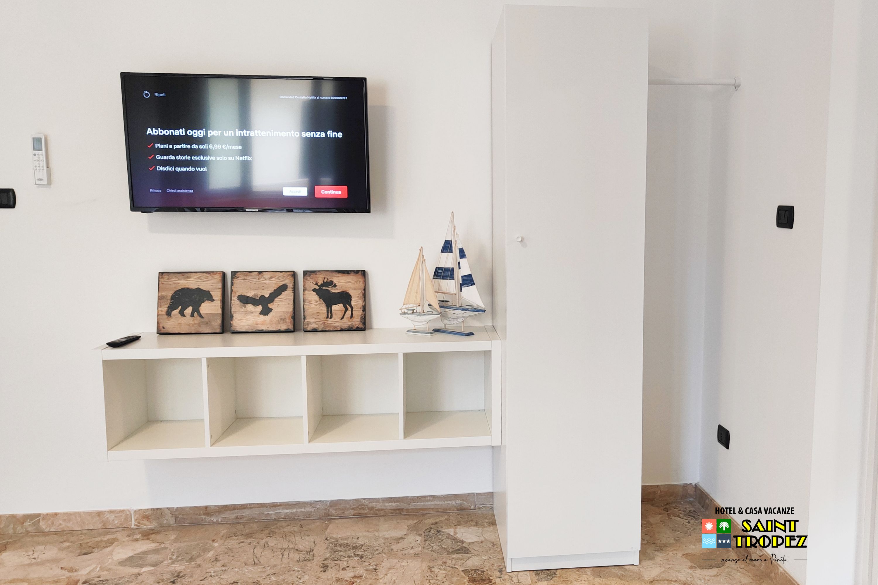 A hotel room with a television mounted on the wall, a remote control on the shelf, and a decorative sailboat.