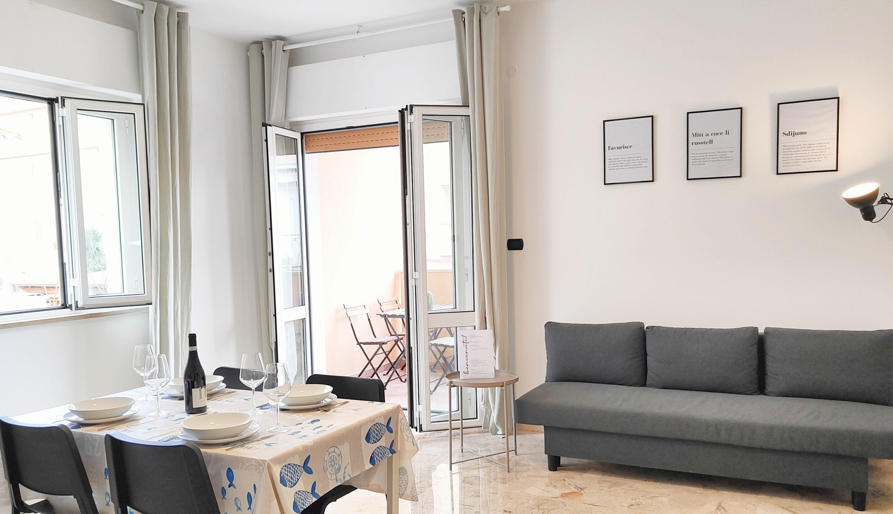 Una sala de estar moderna con un sofá gris, una mesa de comedor con platos y copas de vino, y una pequeña mesita auxiliar.