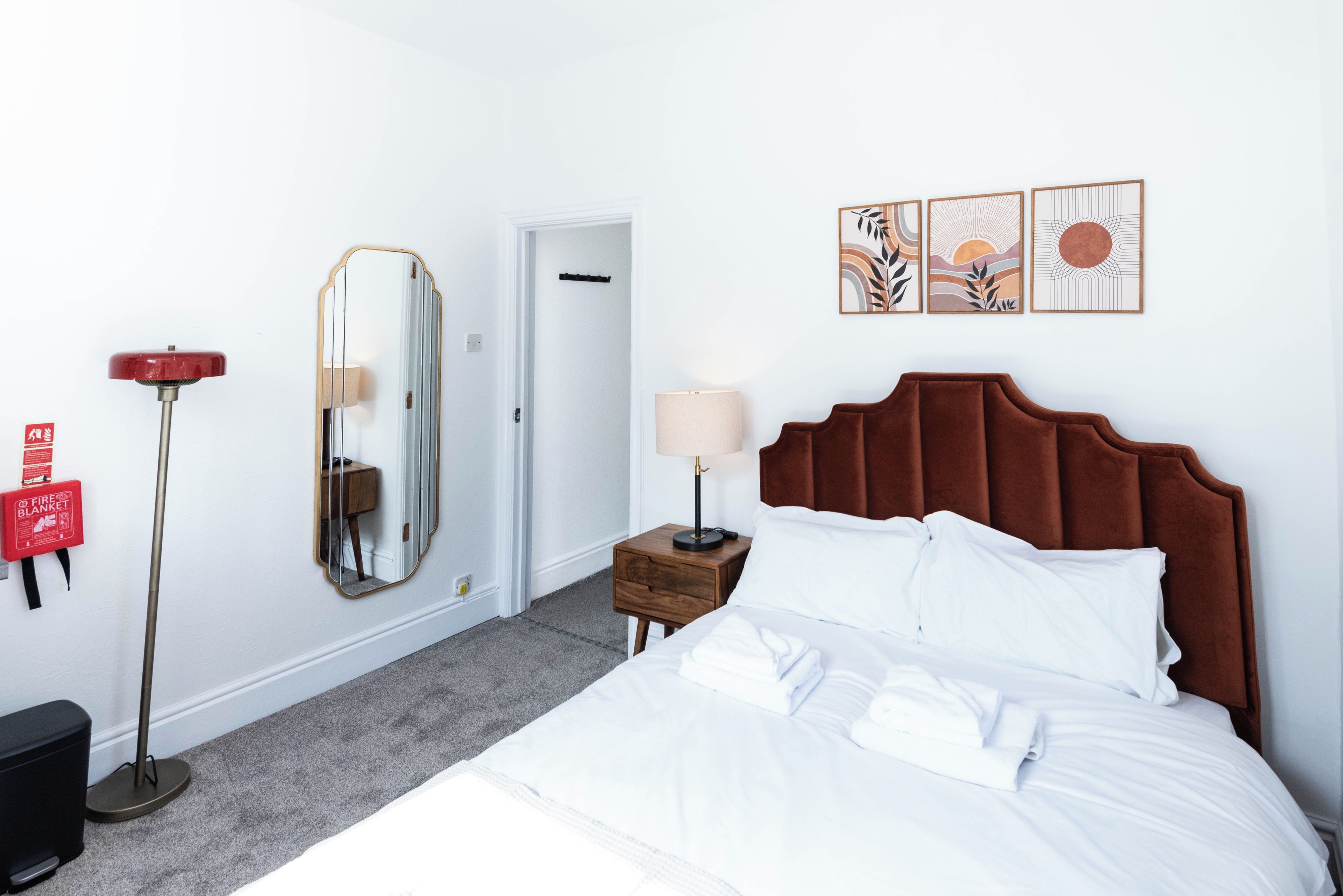 A hotel room with a bed, a bedside table, a lamp, a mirror, a fire blanket, and artwork on the wall.