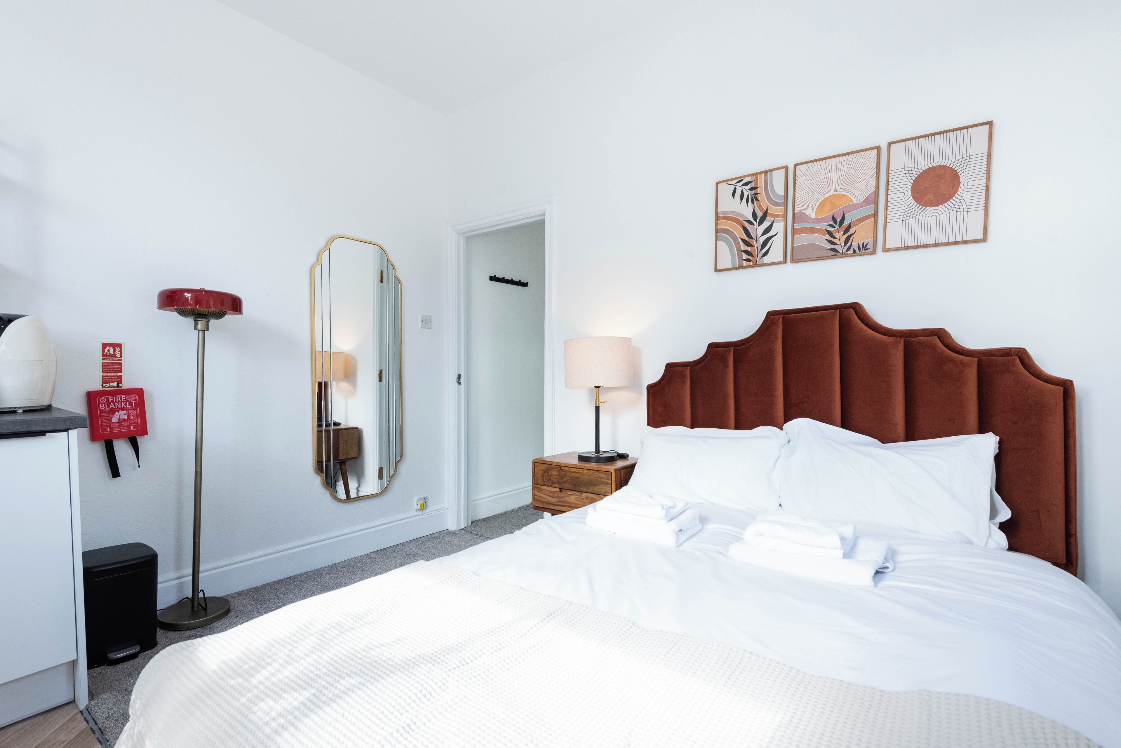 A hotel room with a bed, a bedside table with a lamp, a mirror, a fire blanket, and a red fire alarm.