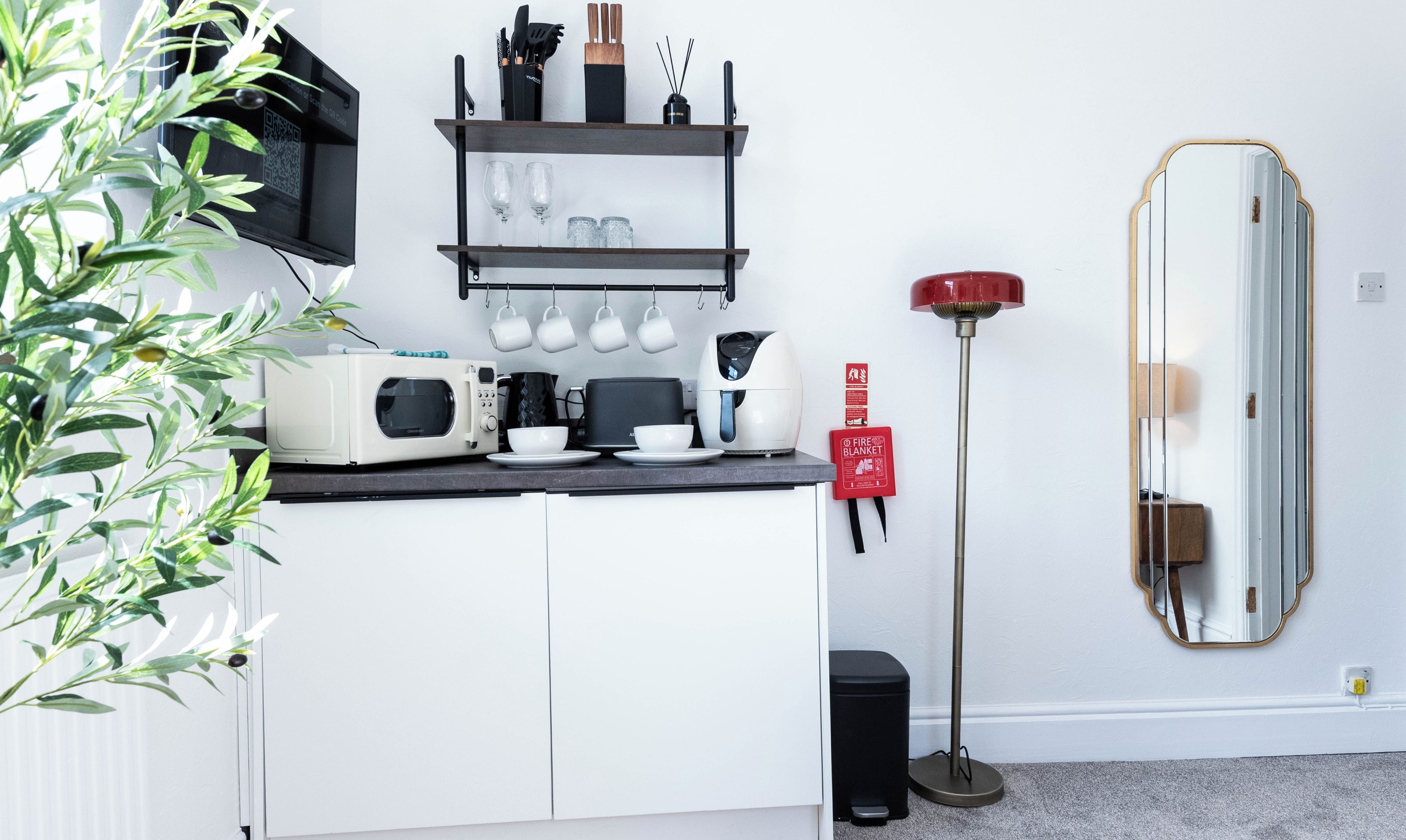 Una cocina moderna con microondas, tostadora y cafetera sobre la encimera, estante con tazas y utensilios, y una manta anti-incendios roja en la pared.