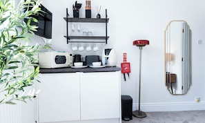 A modern kitchen with a microwave, toaster, and coffee maker on the counter, a shelf with cups and utensils, and a red fire blanket on the wall.