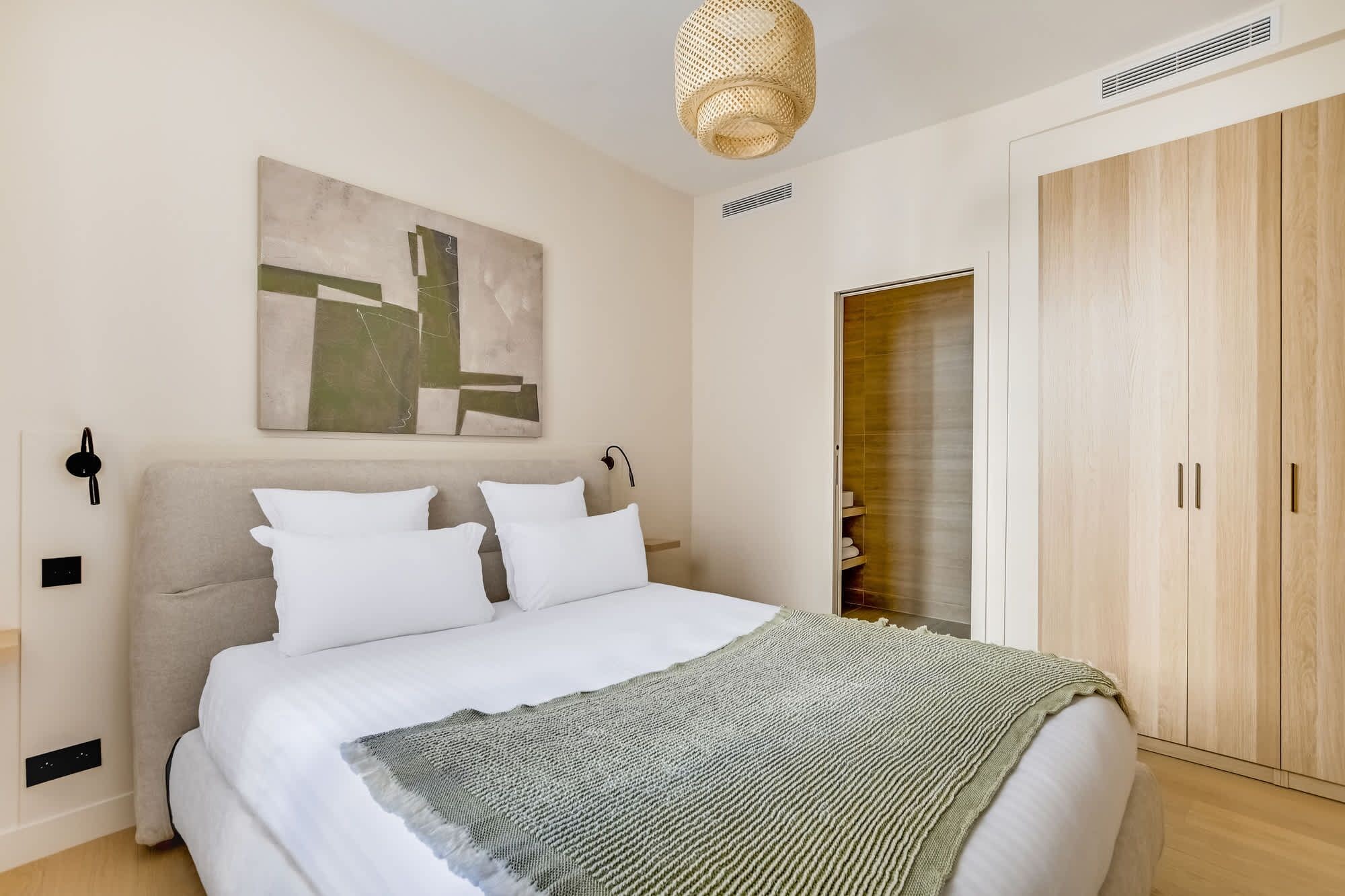 A bedroom with a bed, a wall-mounted abstract painting, a woven light fixture, and a wooden wardrobe.