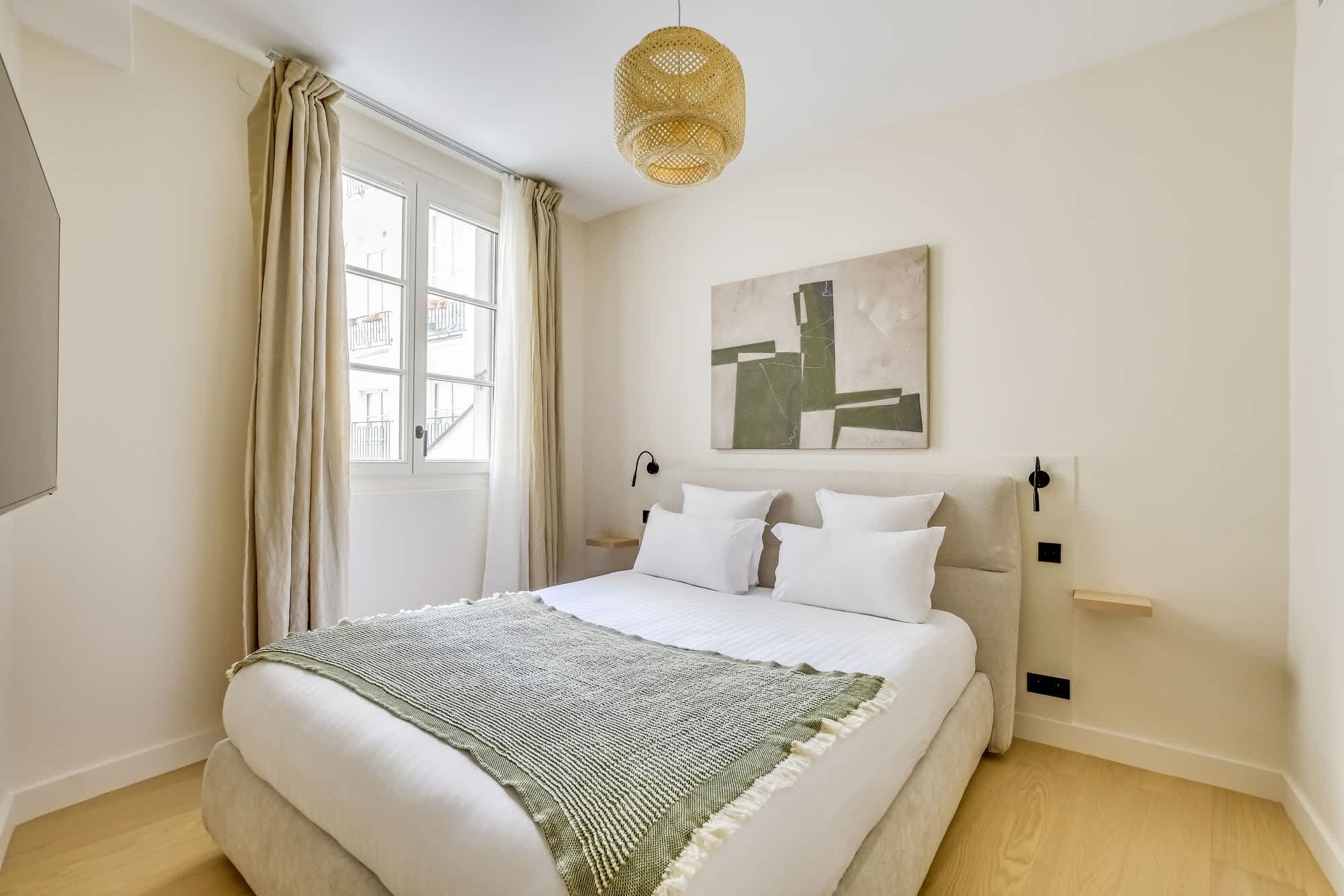 A modern bedroom with a large bed, a window with curtains, a wall-mounted artwork, and a hanging light fixture.