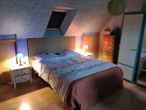 A bedroom with a sloped ceiling, a bed with a patterned bedspread, bedside lamps, a dresser, and a wardrobe. - House with a breathtaking view of the sea – A wild getaway in Brittany (22820 plougrescant)