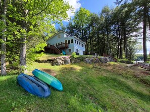 Une maison dotée d’une terrasse, entourée d’arbres et bordée d’une pelouse où se trouvent des kayaks.