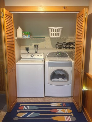 Une buanderie équipée de deux machines à laver, d’un panier et d’une étagère contenant des produits de lessive.
