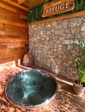 A wooden cabin interior with a stone wall, a sign reading 'SUITE REFUGE', a glass-bottomed hot tub, and a potted plant.